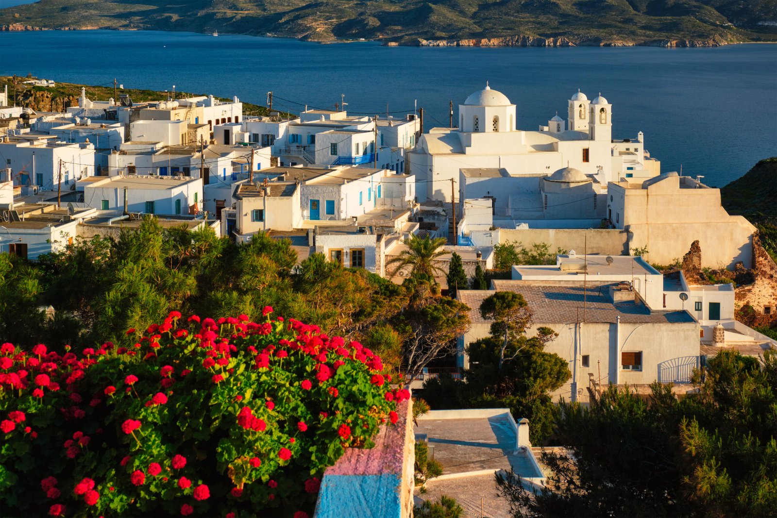 Picturesque scenic view of Greek town Plaka on Milos island over red geranium flowers and Orthodox greek church. Plaka village, Milos island, Greece
